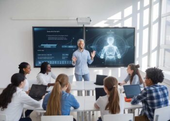 A person is giving a presentation to a group of students in a classroom. Two digital screens display graphics in the background. Students are seated at a table with laptops.