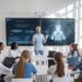 A person is giving a presentation to a group of students in a classroom. Two digital screens display graphics in the background. Students are seated at a table with laptops.