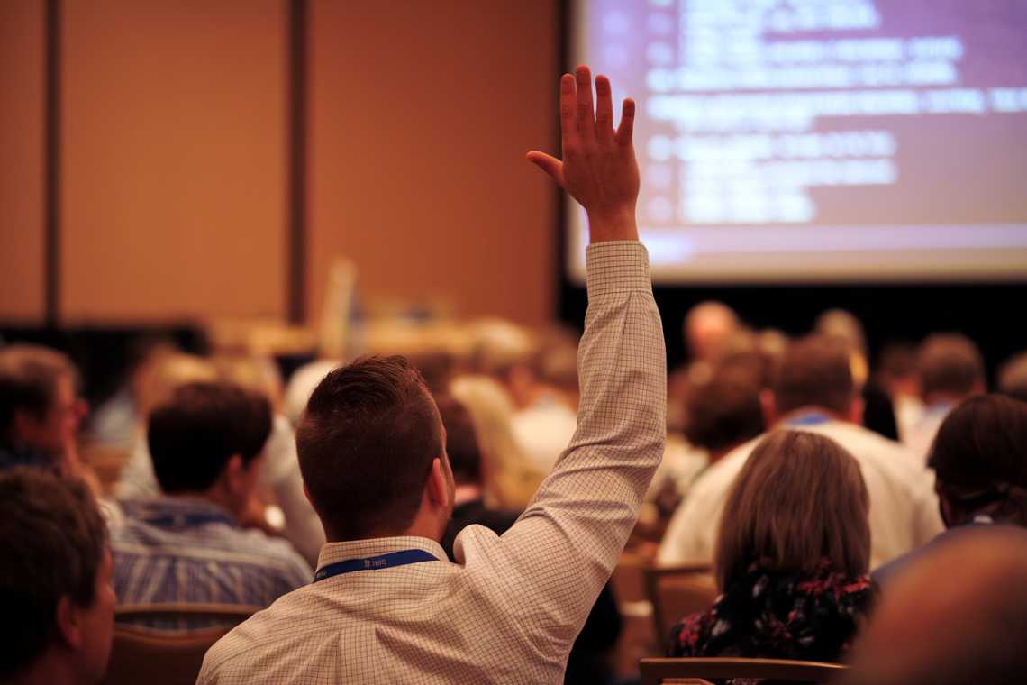 A person raises a hand to ask a question during a conference or seminar attended by a seated audience.