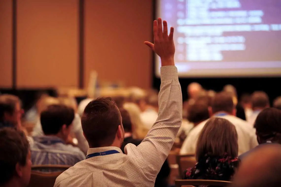 A person raises a hand to ask a question during a conference or seminar attended by a seated audience.