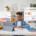 A boy sits at a desk, using a tablet. Various digital graphs and charts are projected around him. Books and stationery are on the desk, and a plant is in the background.