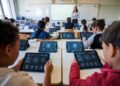 Students sit at desks using tablets in a classroom while a teacher stands near a whiteboard in the background.