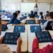 Students sit at desks using tablets in a classroom while a teacher stands near a whiteboard in the background.