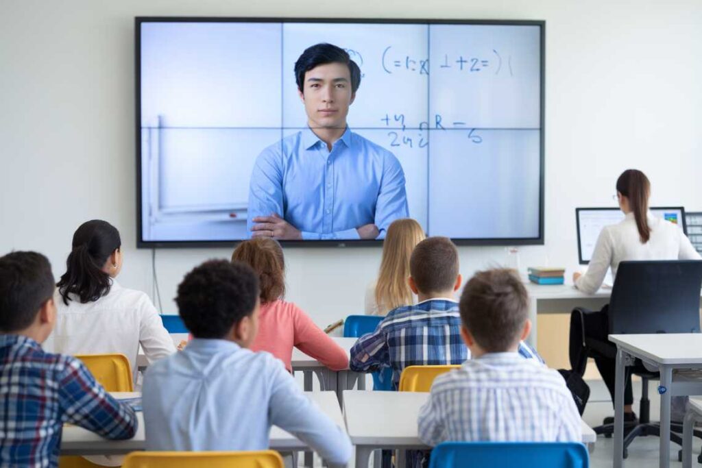 Students sit in a classroom watching a man on a large screen with mathematical equations in the background. A teacher sits at a desk facing the screen.