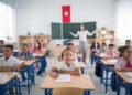 A classroom scene with a diverse group of attentive students sitting at desks. A teacher stands in front of a blackboard, gesturing with arms open. A red flag hangs on the wall.