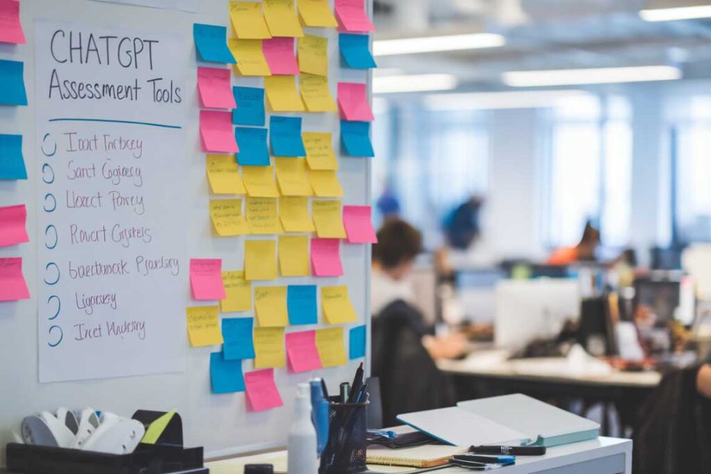 Office scene with a whiteboard labeled "CHATGPT Assessment Tools" covered in sticky notes and an open-plan workspace in the background.