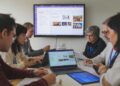 A group of people sitting around a table, engaging in a meeting. Laptops and documents are on the table, and a screen displays information in the background.