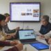 A group of people sitting around a table, engaging in a meeting. Laptops and documents are on the table, and a screen displays information in the background.