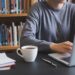 Using Grammarly to Improve Student Writing: Enhancing Academic Performance Person typing on a laptop at a black table with a cup of coffee, a notebook, and a pen, in front of bookshelves.