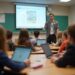 A teacher stands at the front of a classroom, gesturing while students sit at desks with laptops, focused on a presentation displayed on a screen.