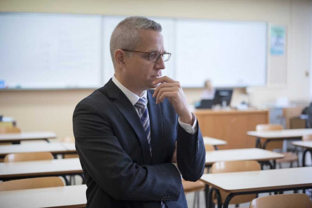 Man in a suit and glasses standing thoughtfully in an empty classroom with desks and a whiteboard.