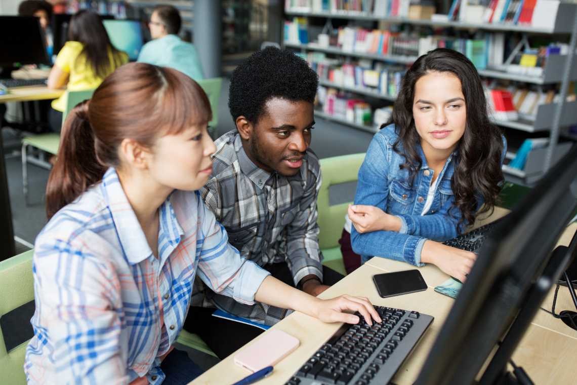 Three people are seated at a computer in a library, focusing on the screen. Books are visible in the background.